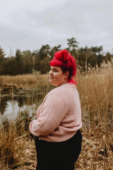 Lisa Jansen staat in een veld en kijkt dromerig weg van de camera. Ze heeft rood haar en draagt een lichtroze trui en een zwarte broek.