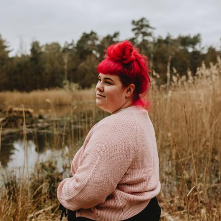 Lisa Jansen staat in een veld en kijkt dromerig weg van de camera. Ze heeft rood haar en draagt een lichtroze trui en een zwarte broek.